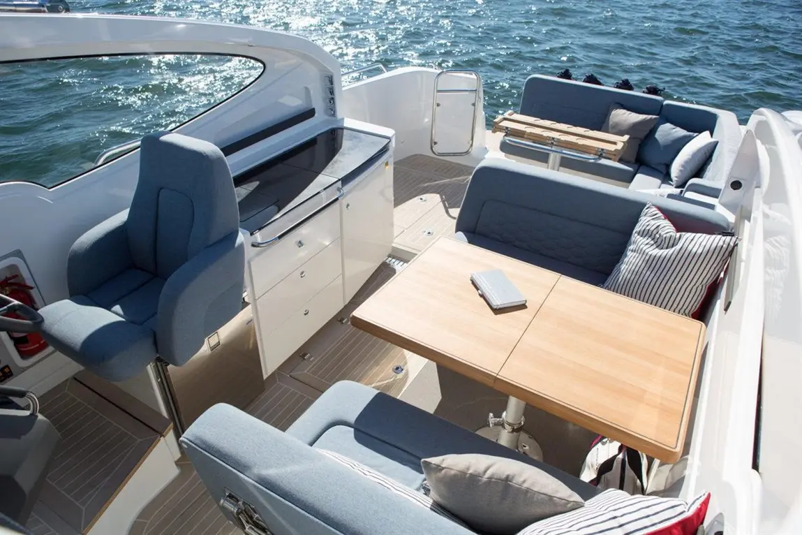 Interior of a modern boat featuring a wooden table, grey seating with cushions, and a steering area overlooking water.